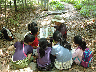 방과후학교 수업 장면-숲속에서 아이들이 모여 앉아 선생님이 보여주는 책을 보고 있는 모습