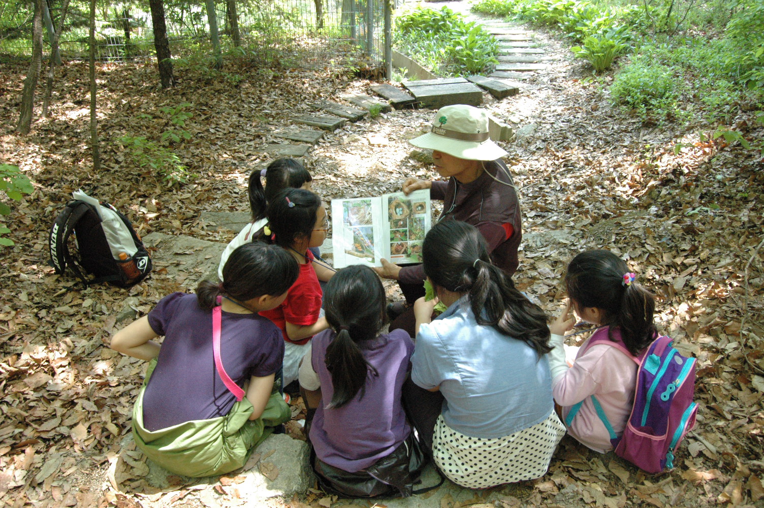방과후학교 수업 장면-숲속에서 아이들이 모여 앉아 선생님이 보여주는 책을 보고 있는 모습